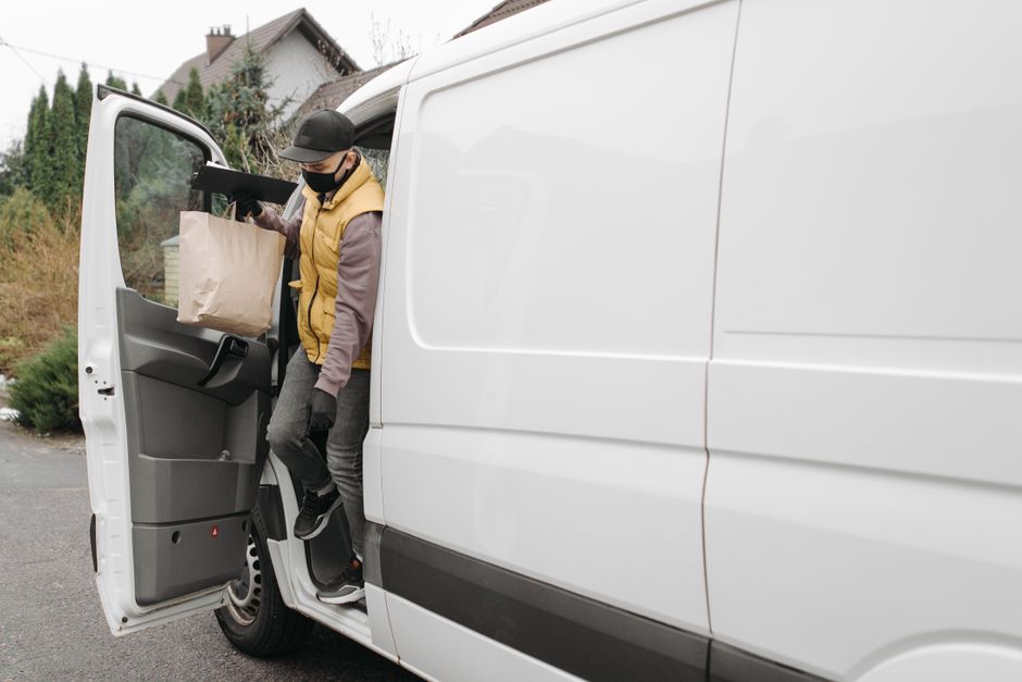 A person wearing a black face mask, grey cap, yellow padded vest over a grey long-sleeve shirt, and black gloves is seen stepping out of a white cargo van, holding a brown paper bag in their left hand